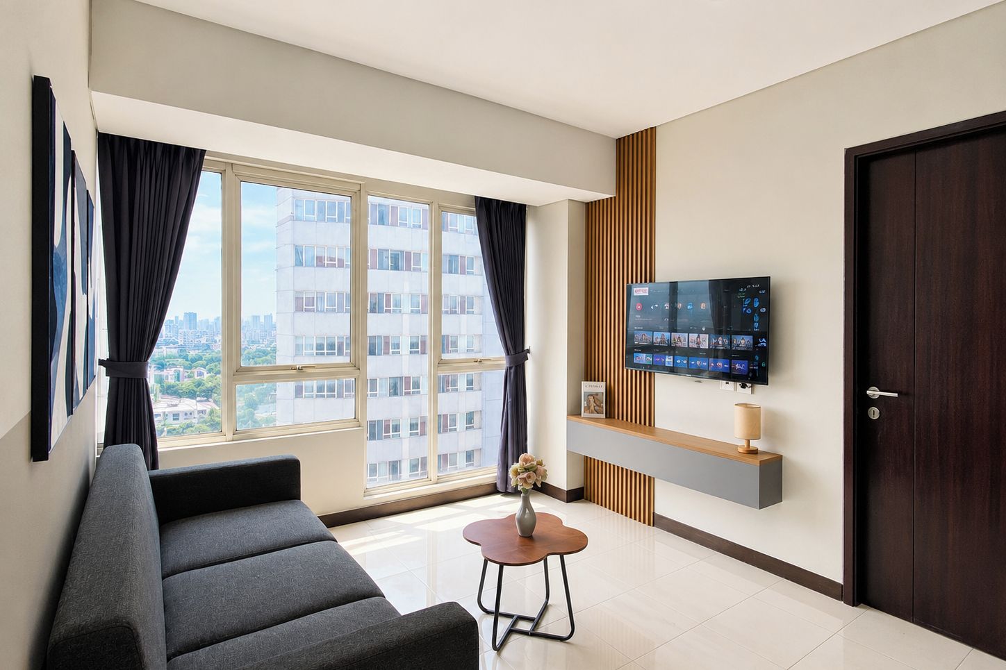 A modern living room with a sofa, a small table, a TV mounted on the wall, and a large window with curtains.