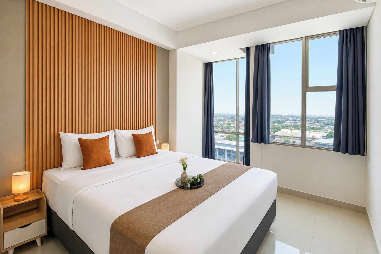 Una habitación de hotel con una cama grande, un cabecero de madera, una mesita de noche, una lámpara de noche y vistas de la ciudad a través de amplias ventanas.