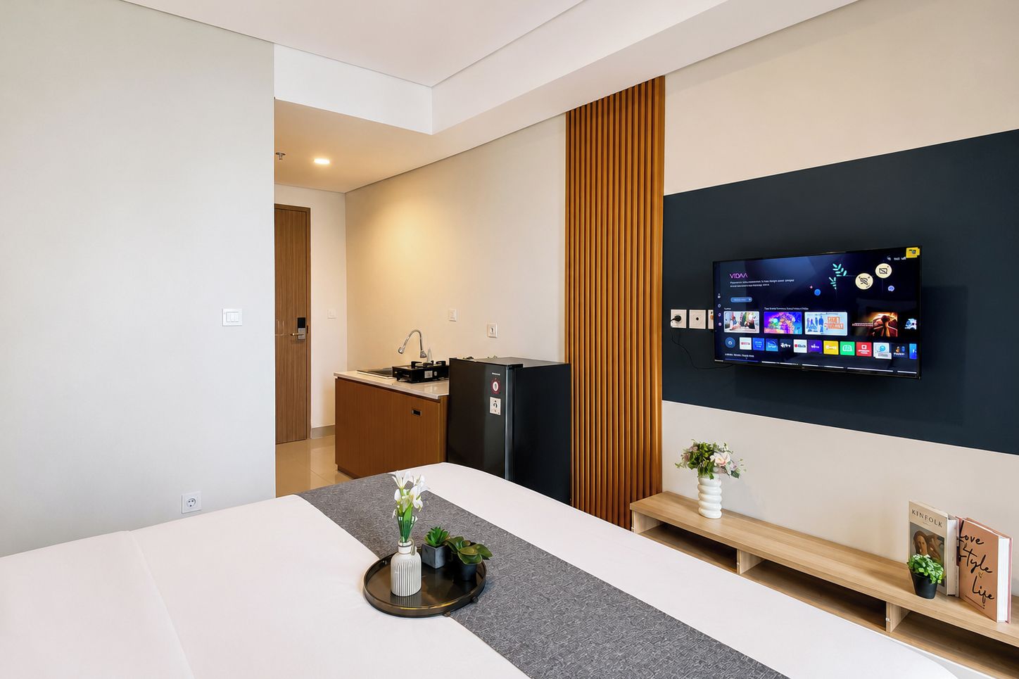 Una sala moderna con un televisor de pantalla plana montado en la pared, un mueble de madera, un refrigerador y un comedor con mesa y banco.