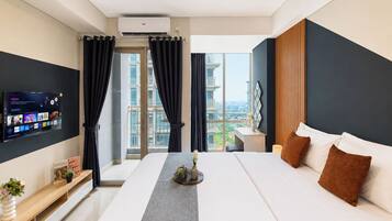 Habitación de hotel con una cama grande, televisor montado en la pared, balcón con cortinas y vistas a la ciudad.