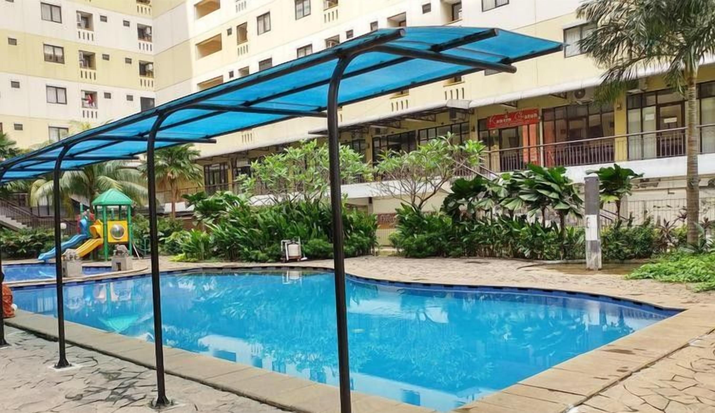 Una piscina amb una zona coberta, una estructura de jocs infantils i un edifici amb balcons al fons.