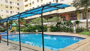 A swimming pool with a covered area, a children's play structure, and a building with balconies in the background. - Lovely And Cozy Studio At Apartment Kebagusan City (Jakarta)