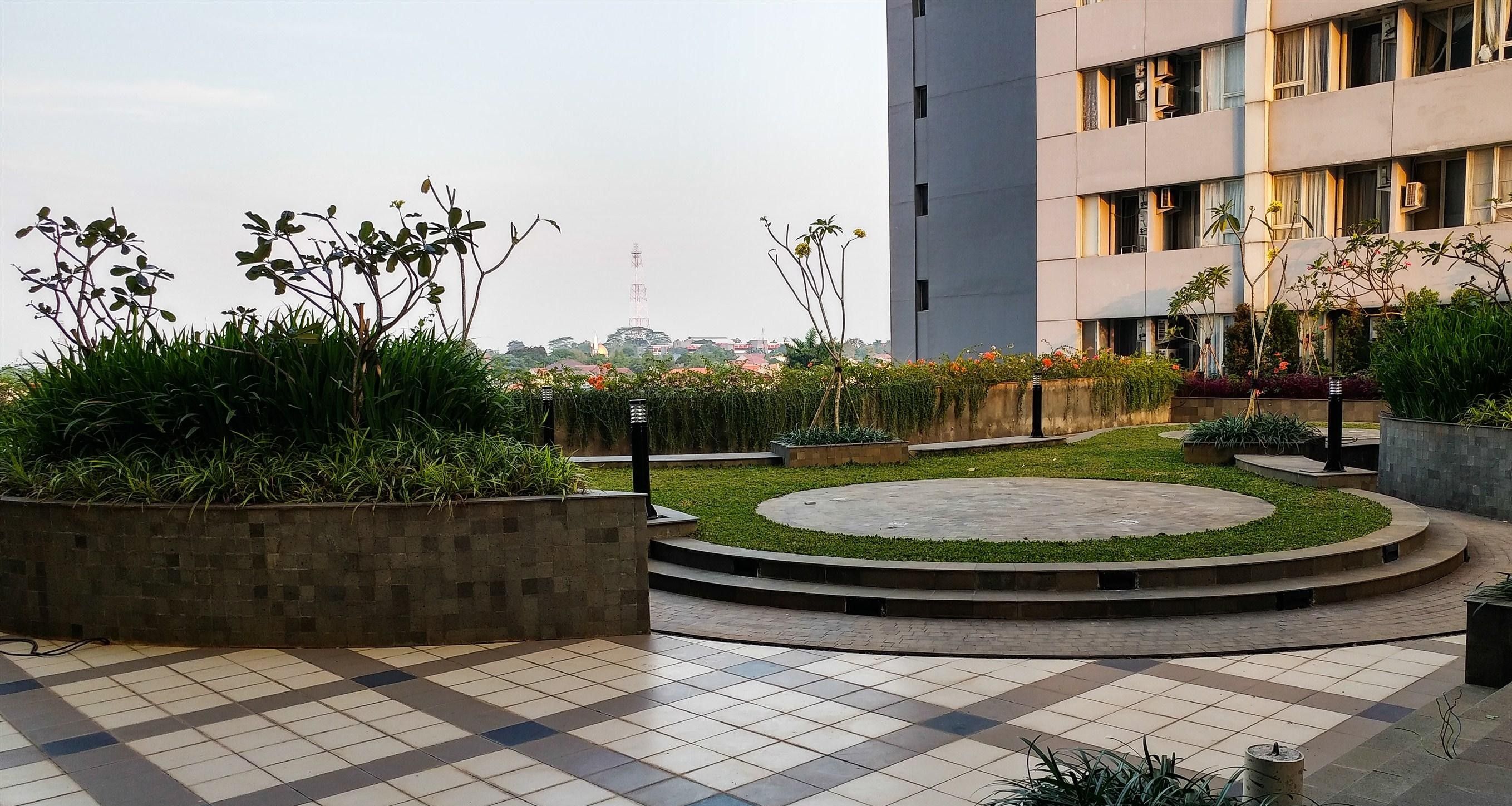 A rooftop garden with a circular seating area, surrounded by planters with various plants, and a view of a cityscape in the background.