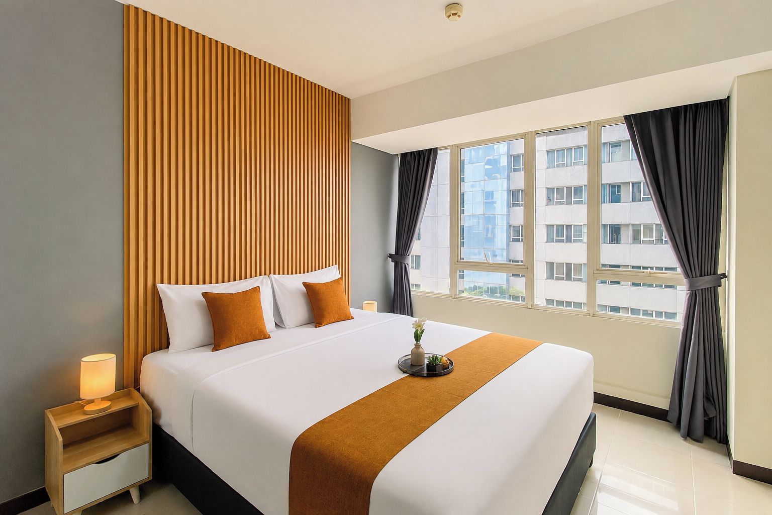 A hotel room with a large bed, a nightstand, a window with curtains, and a view of buildings outside.