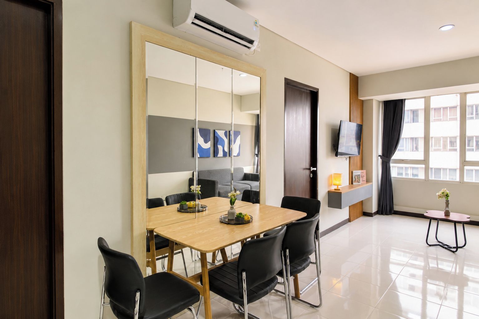 A hotel room with a wooden dining table, black chairs, a large mirror, a flat-screen TV mounted on the wall, and a window with curtains.