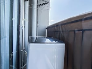 Eine kompakte, weiße Waschmaschine steht in einer Ecke des Raumes, neben einer Glastür und einem Metalldrahtgitter.