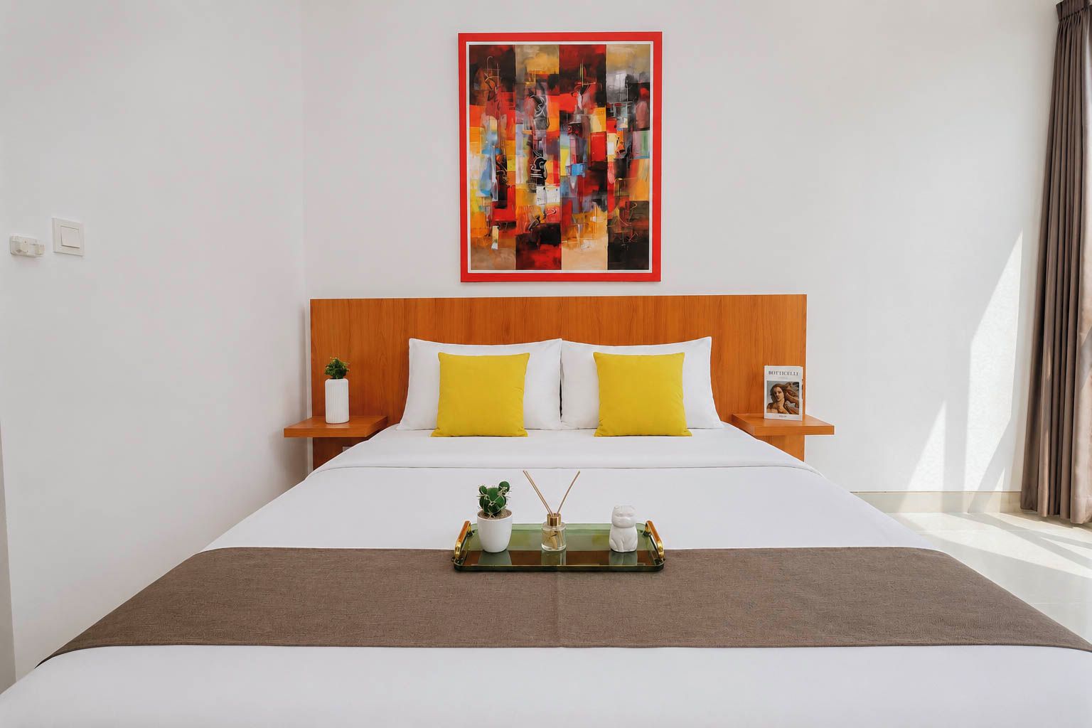 Habitación de hotel con una cama grande, cabecero de madera, ropa de cama blanca, dos almohadas amarillas, una mesita de noche con una planta, un cuadro enmarcado y una bandeja con una planta y una vela.