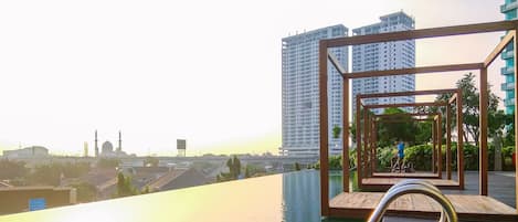 A rooftop infinity pool with a wooden deck and metal railings, overlooking a cityscape with high-rise buildings and a mosque.