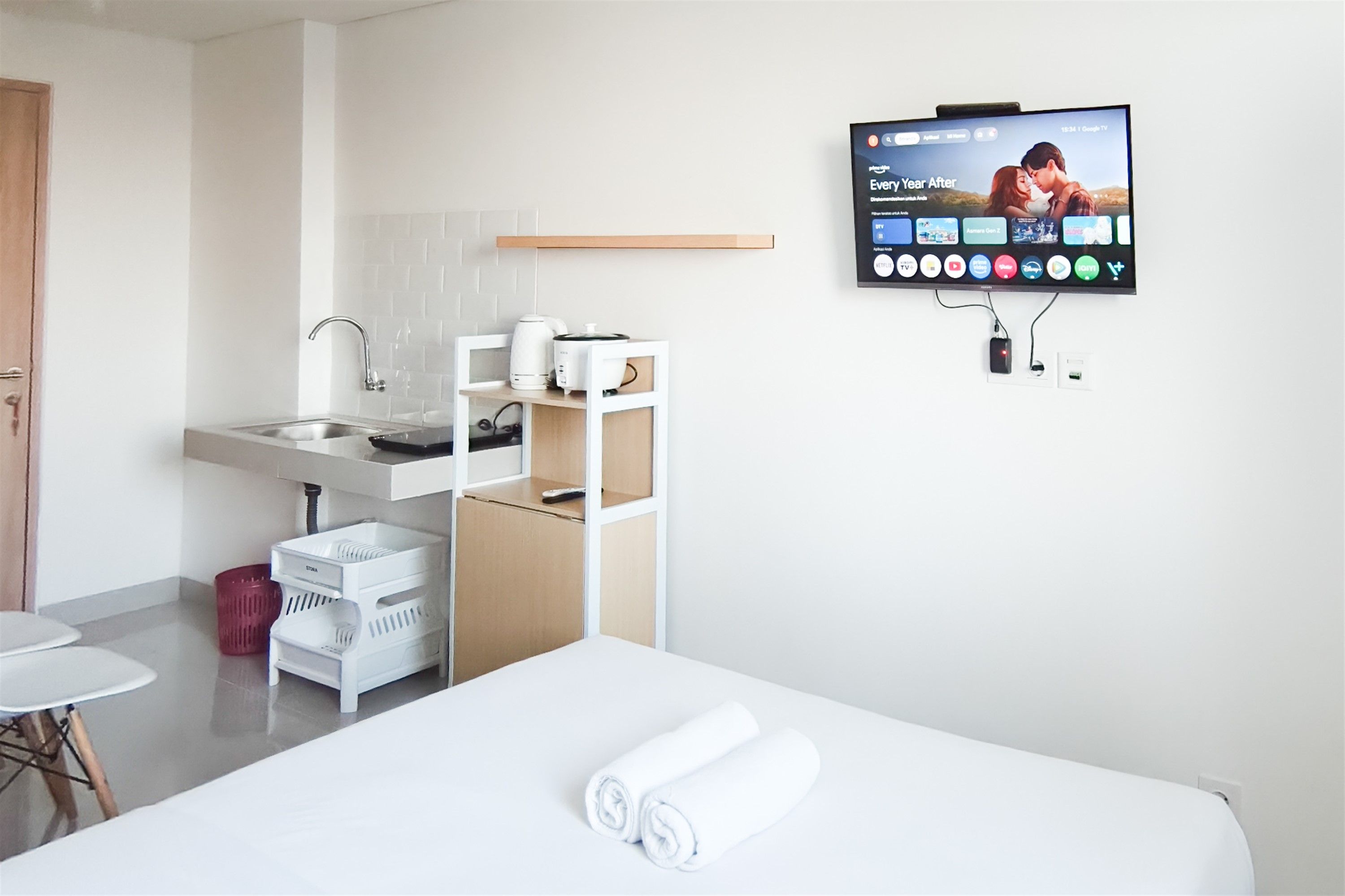 A hotel room with a bed, a TV mounted on the wall displaying a movie, a kitchen area with a sink and a coffee maker, and a small table with a red basket.
