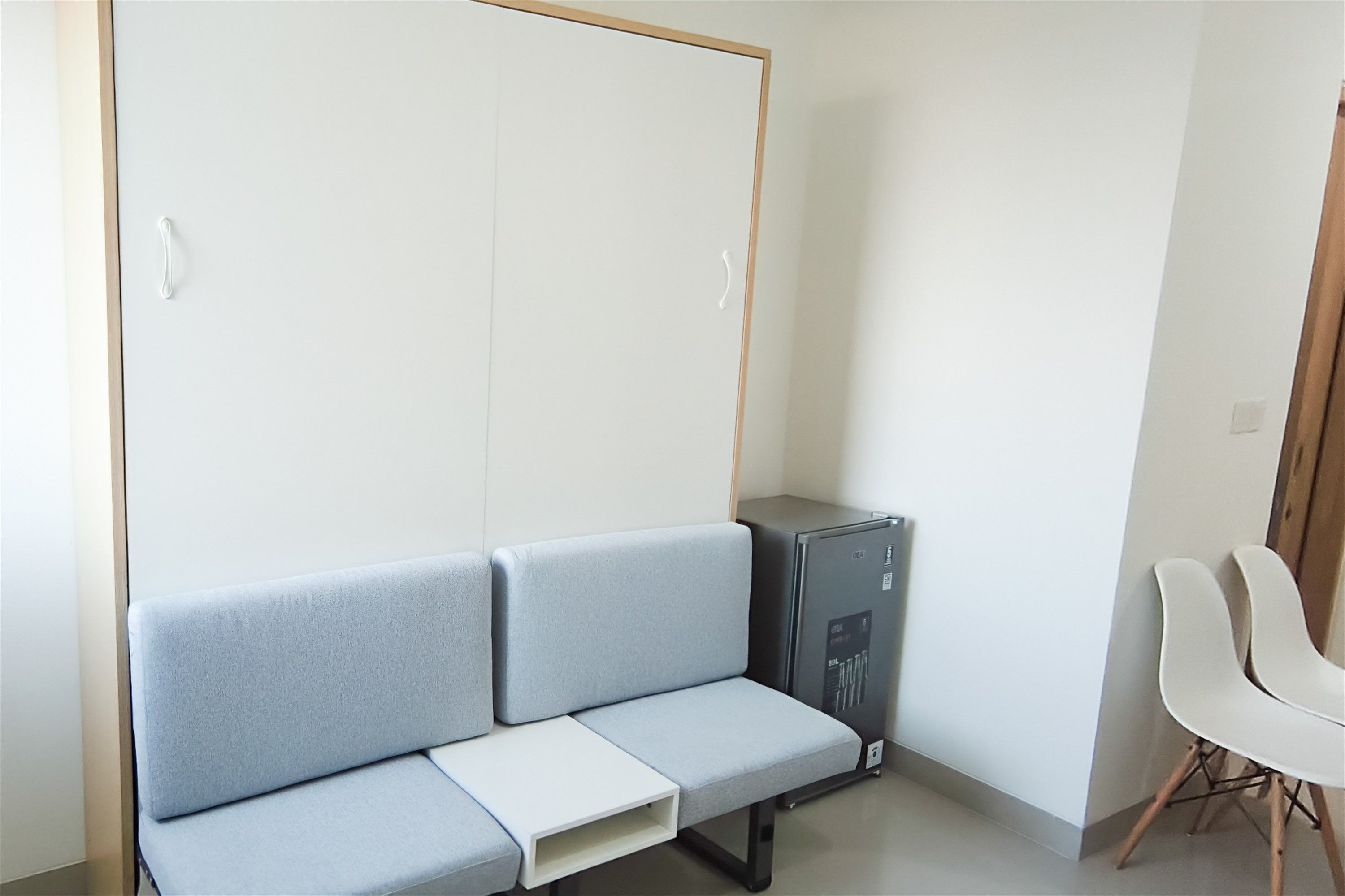 Una habitación con un sofá gris, una mesita blanca, un armario negro y una silla blanca.