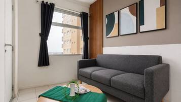 Una sala moderna con un sofá gris, una mesita redonda de madera con un mantel verde, un florero blanco con una planta y una ventana con cortinas negras.