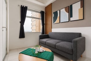 Un salon moderne avec un canapé gris, une petite table ronde en bois recouverte d’une nappe verte, un vase blanc contenant une plante et une fenêtre ornée de rideaux noirs.