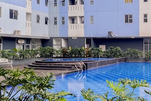 Une piscine accessible par des marches, entourée de verdure et bordée d’un immeuble à plusieurs étages doté de balcons.