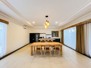 A spacious dining area with a long wooden table and chairs, a kitchen in the background, and large windows with curtains. - 3Br Moriva Villa Ungasan Near Melasti Beach (Jimbaran)