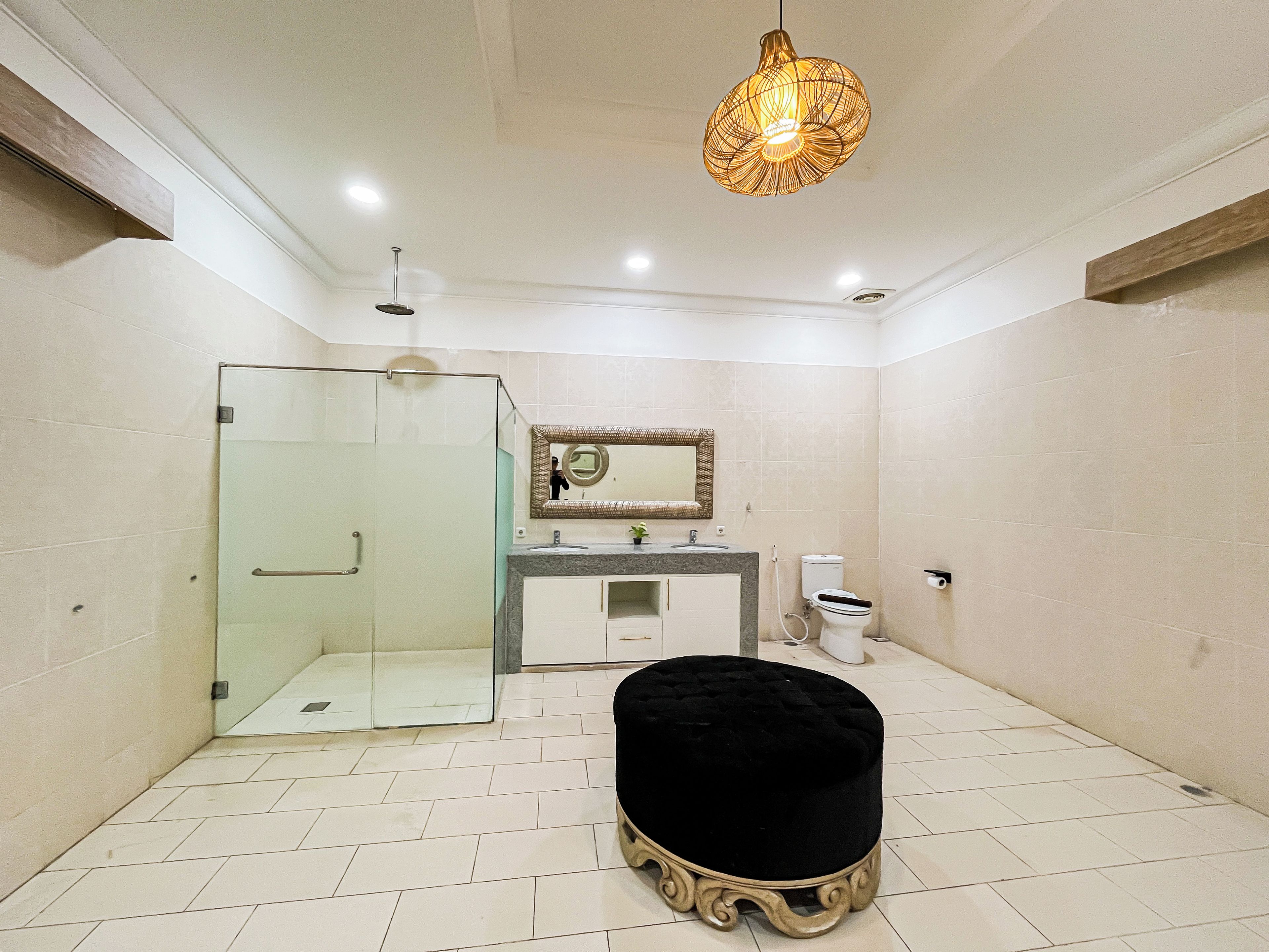 A bathroom with a glass shower enclosure, a vanity with a mirror, a toilet, and a black ottoman.