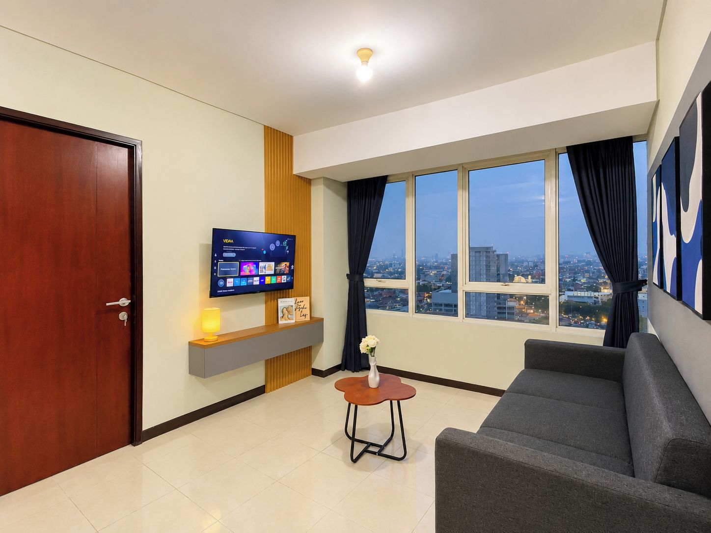 A modern living room with a sofa, a TV mounted on the wall, a small table with a vase, and a city view through large windows.