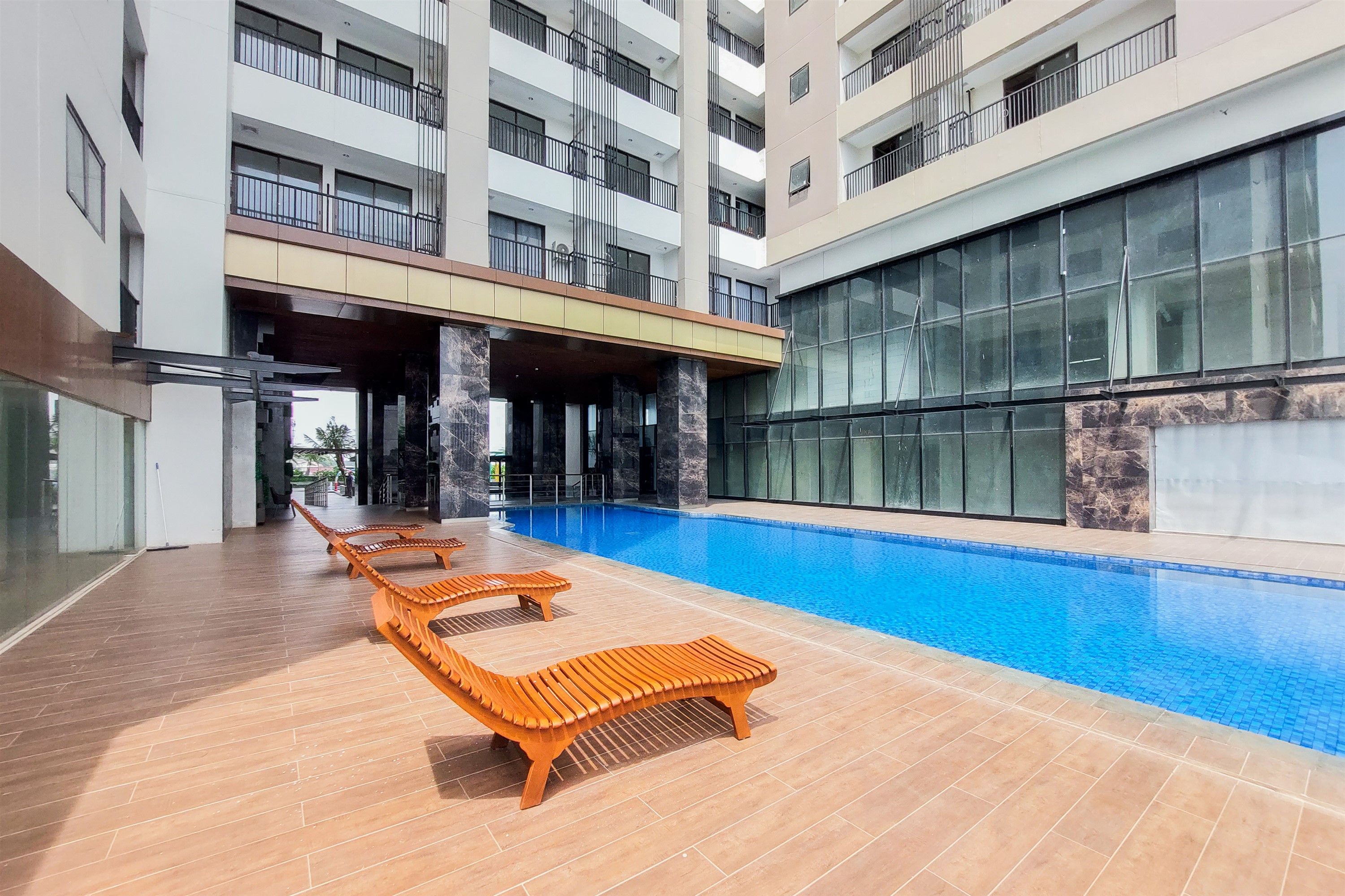 A modern hotel with a swimming pool, wooden lounge chairs, and a multi-story building with balconies.