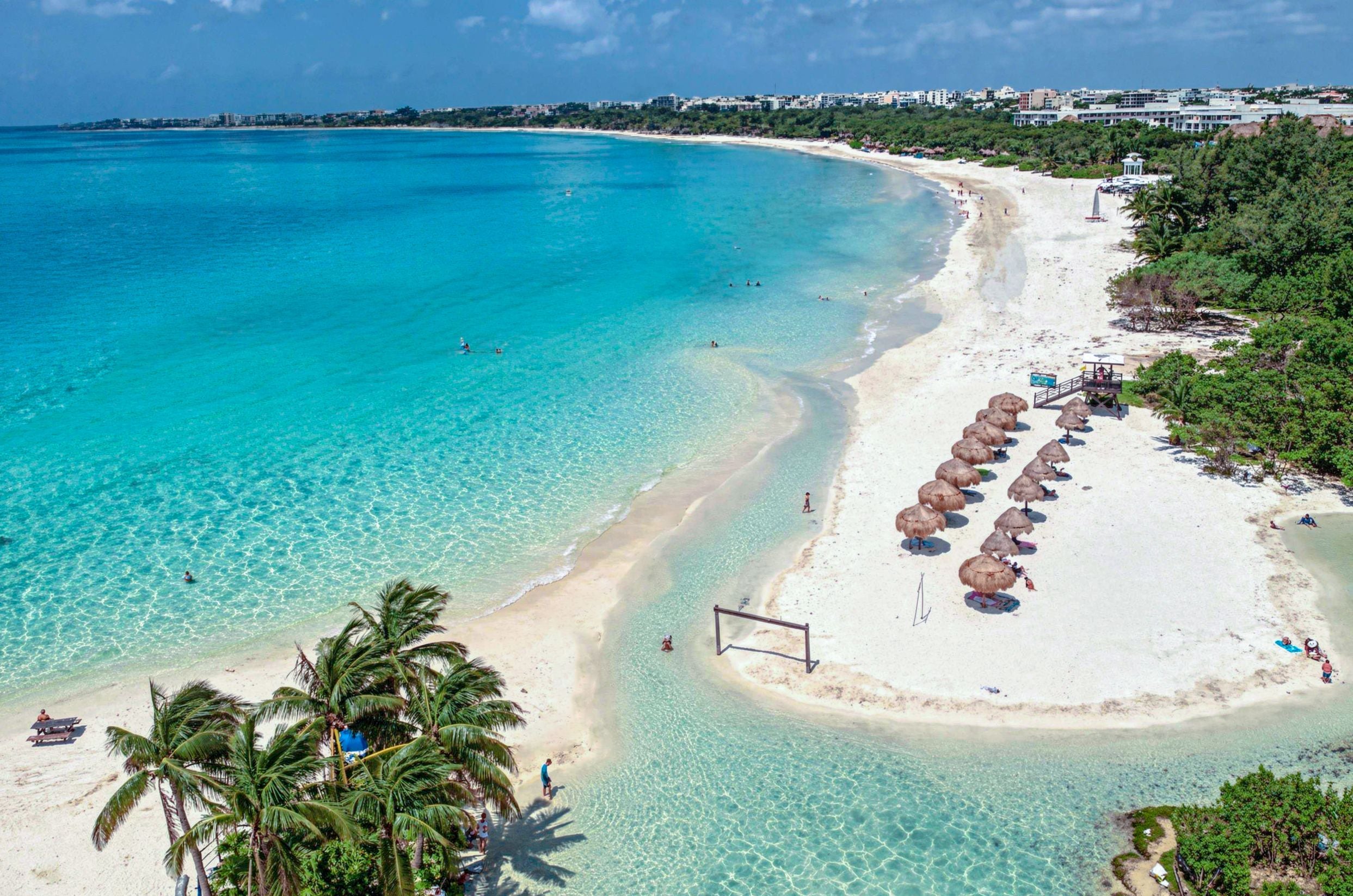 Une plage aux eaux turquoise et limpides, un rivage sablonneux et une rangée de parasols à toits de chaume.