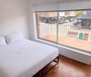 A hotel room with a bed, a window with blinds, and a view of the street outside. - Bondi 1BR Apartment, Private Balcony | 2 Min Walk to Beach, Sleeps 4 (Bondi Beach)