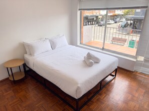 A single-bed room with a wooden floor, a small round table, and a window with a view of the street. - Bondi 1BR Apartment, Private Balcony | 2 Min Walk to Beach, Sleeps 4 (Bondi Beach)