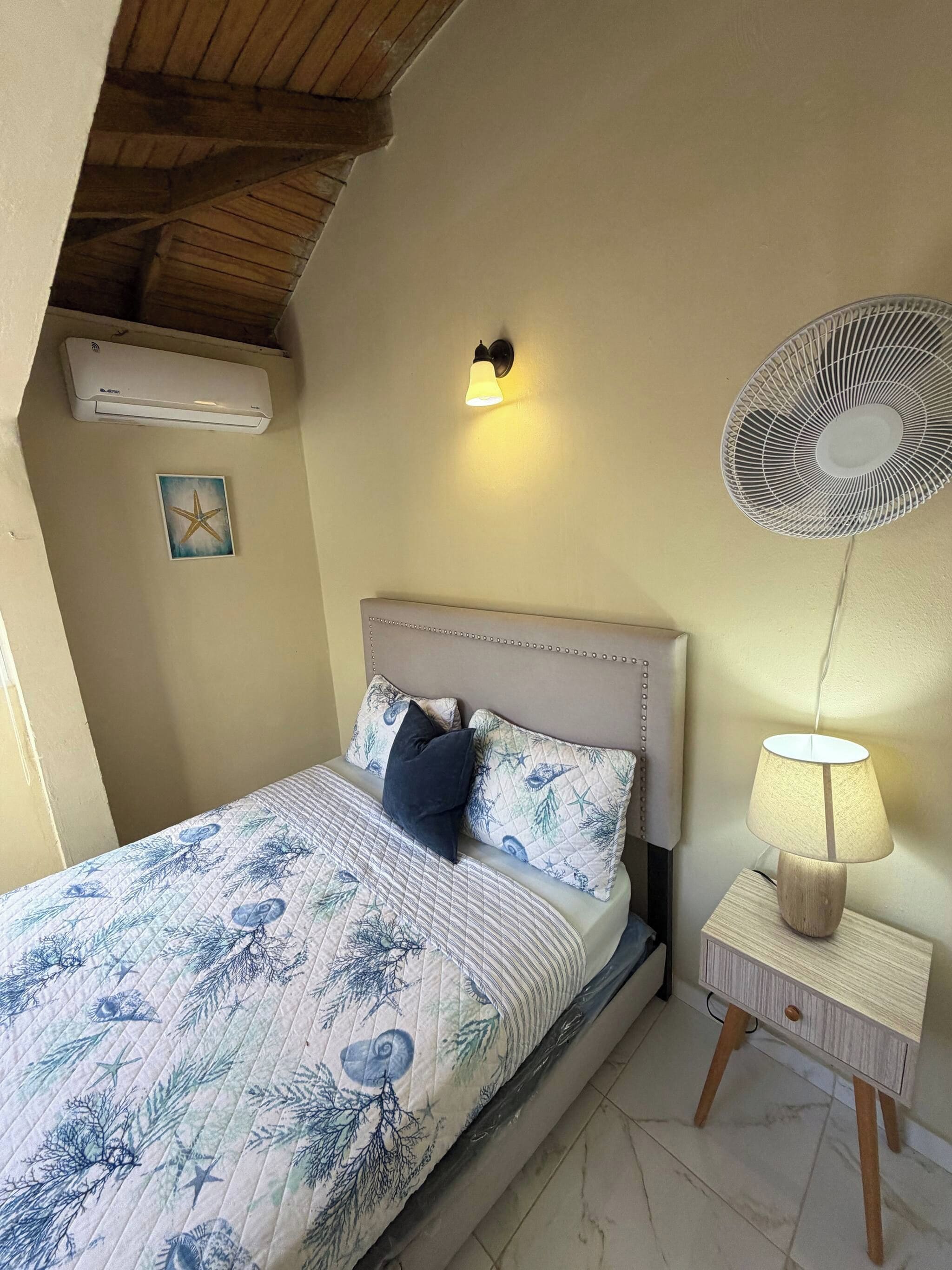 A bedroom with a bed, a wall-mounted fan, a bedside lamp, and an air conditioning unit.