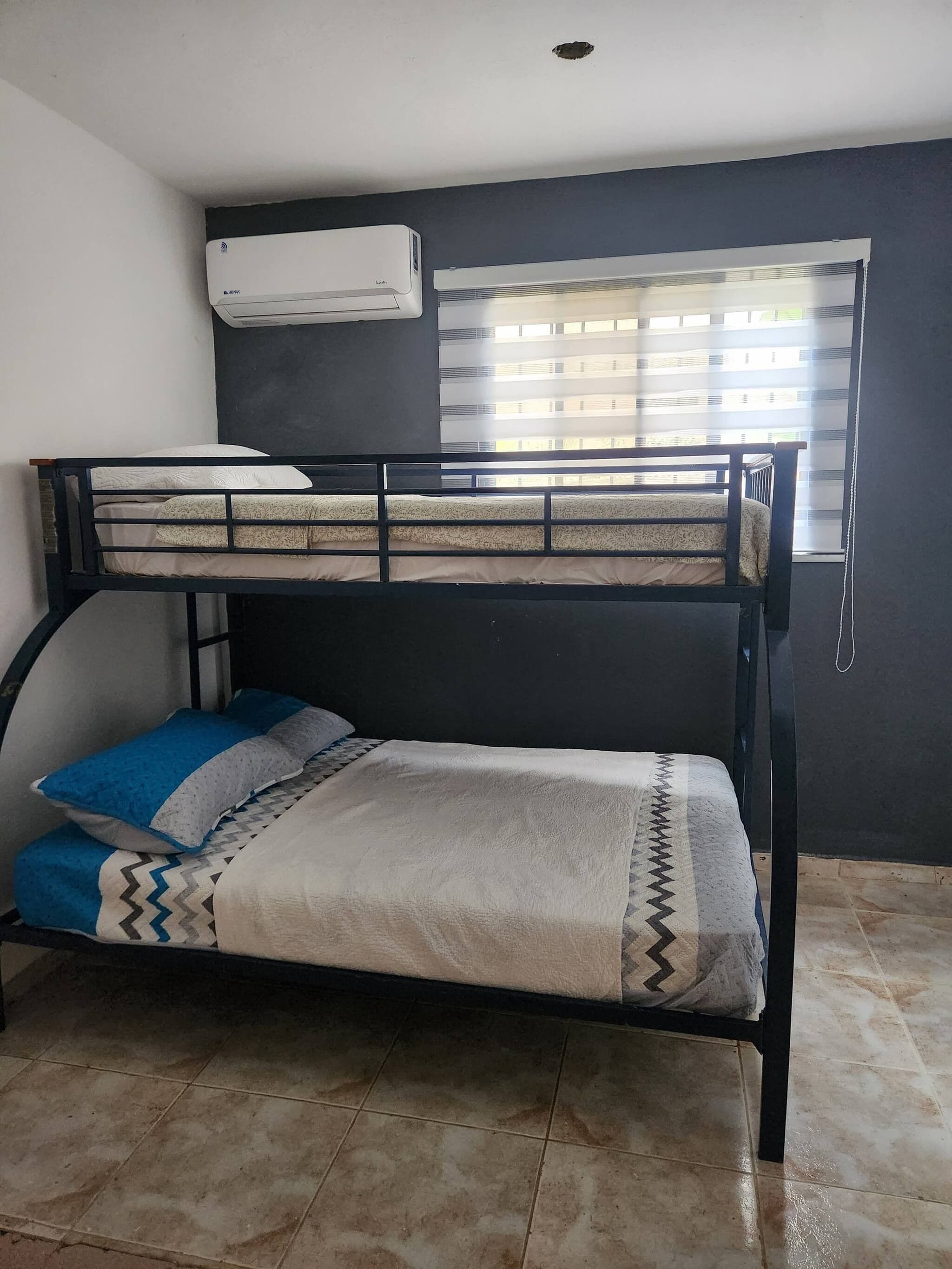 A bunk bed with a mattress, pillows, and a blanket. There is a window with blinds and an air conditioning unit mounted on the wall.