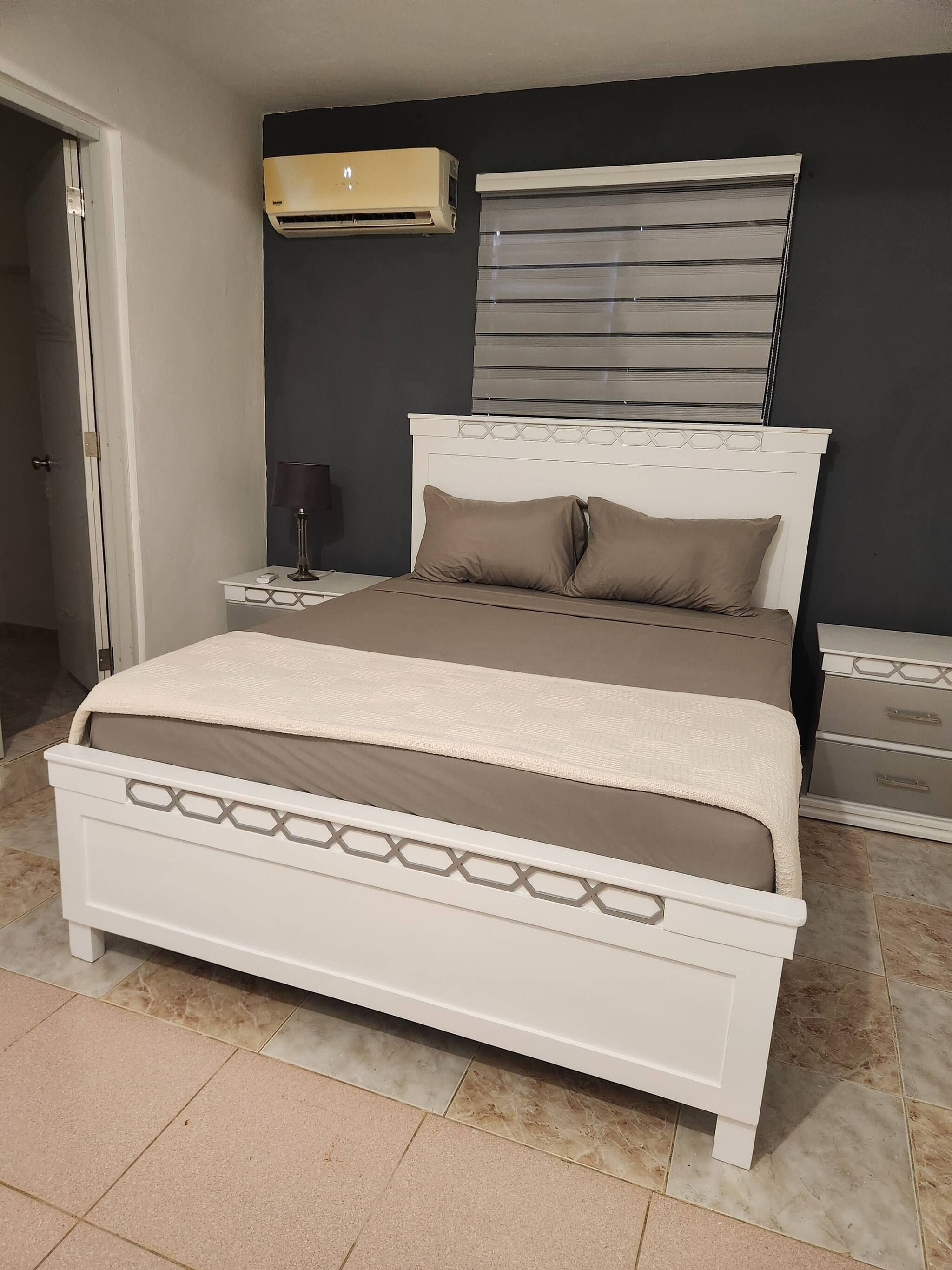 Un dormitorio con cama, mesitas de noche, aire acondicionado y ventana con persianas.