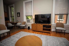 Ett vardagsrum med en platt-TV placerad på ett träskåp, två fåtöljer, ett soffbord och ett stort fönster med persienner.