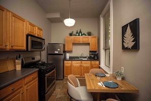 A compact kitchen with wooden cabinets, a stainless steel refrigerator, a microwave, and a gas stove. There is a dining table with chairs and a window with blinds. - Newly furnished 2BR Apt-Walkable + Near hospitals & universities + Ample Parking (Cincinnati)