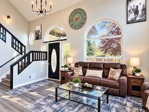 A spacious living room with a brown leather sofa, a glass coffee table, and a large window with a view of trees. - Massive Family Getaway with private apartment, hot tub and pool table. (Bend)