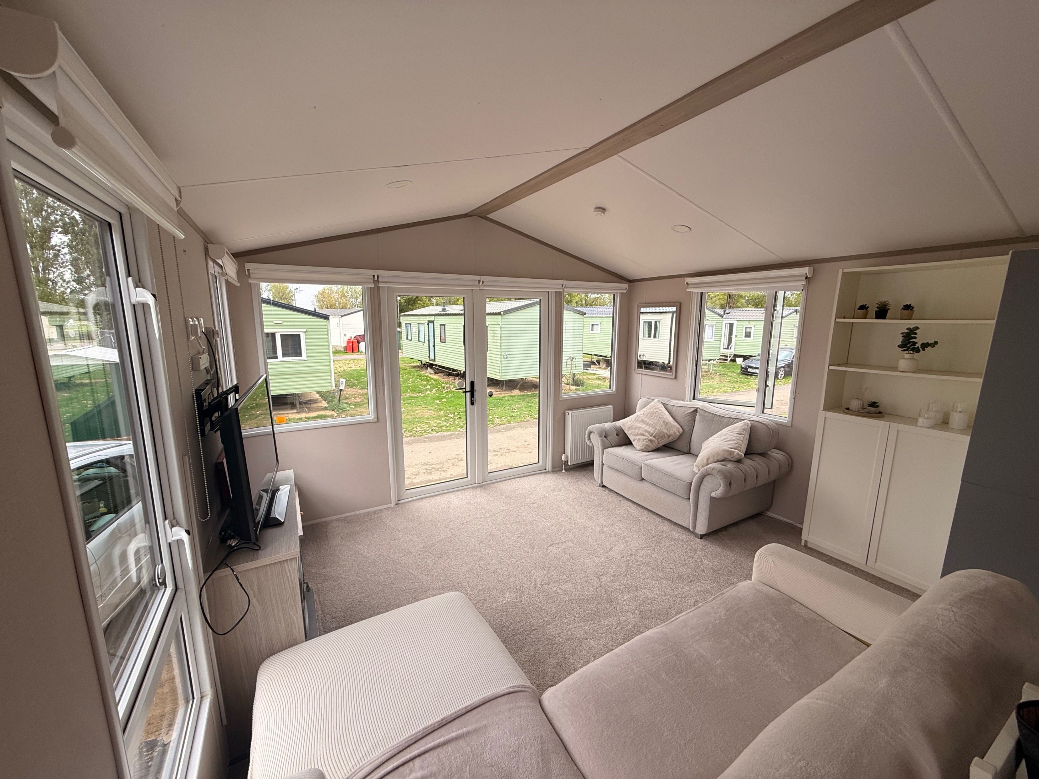 A room with a sofa, a small table, a cabinet with shelves, and a sliding glass door leading to an outdoor area with mobile homes.