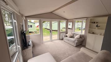 A room with a sofa, a small table, a cabinet with shelves, and a sliding glass door leading to an outdoor area with mobile homes.