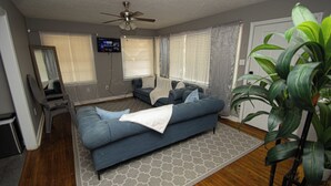 Ein Wohnzimmer mit einem blauen Sofa, einem Deckenventilator, einem an der Wand befestigten Fernseher und einer großen Pflanze.