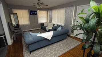 Ein Wohnzimmer mit einem blauen Sofa, einem Deckenventilator, einem an der Wand befestigten Fernseher und einer großen Pflanze.