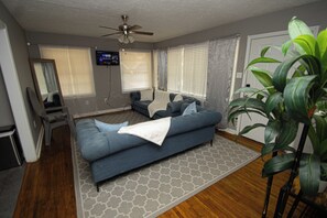 A living room with a blue sofa, a ceiling fan, a television on the wall, and a large plant. - House Sleeps 8 with Air Conditioning (North Little Rock)
