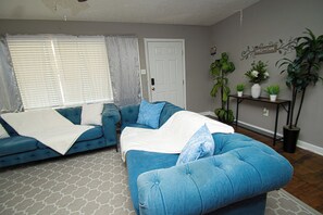 A living room with a blue sofa set, a white door, and a plant shelf. - House Sleeps 8 with Air Conditioning (North Little Rock)