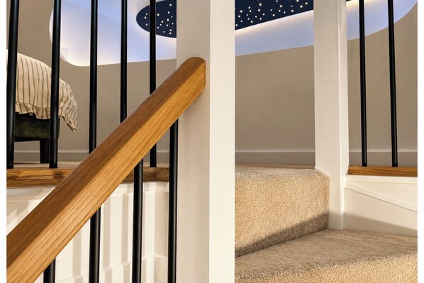 Una escalera con barandillas de madera y balaustres negros, escalones alfombrados y vista a una habitación con una proyección de cielo estrellado en la pared.