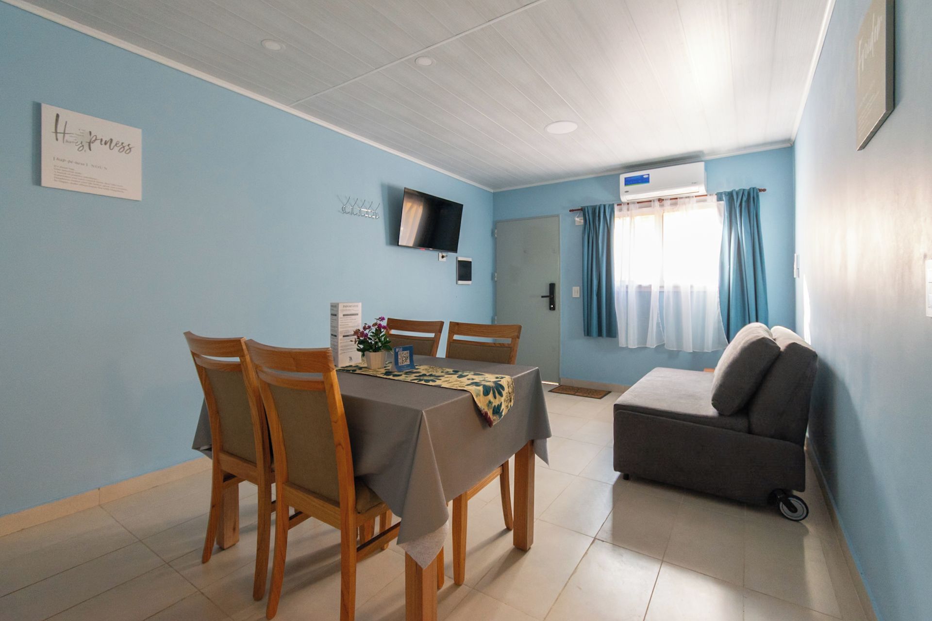 Un comedor con mesa y sillas, un sofá, un televisor en la pared y una ventana con cortinas.