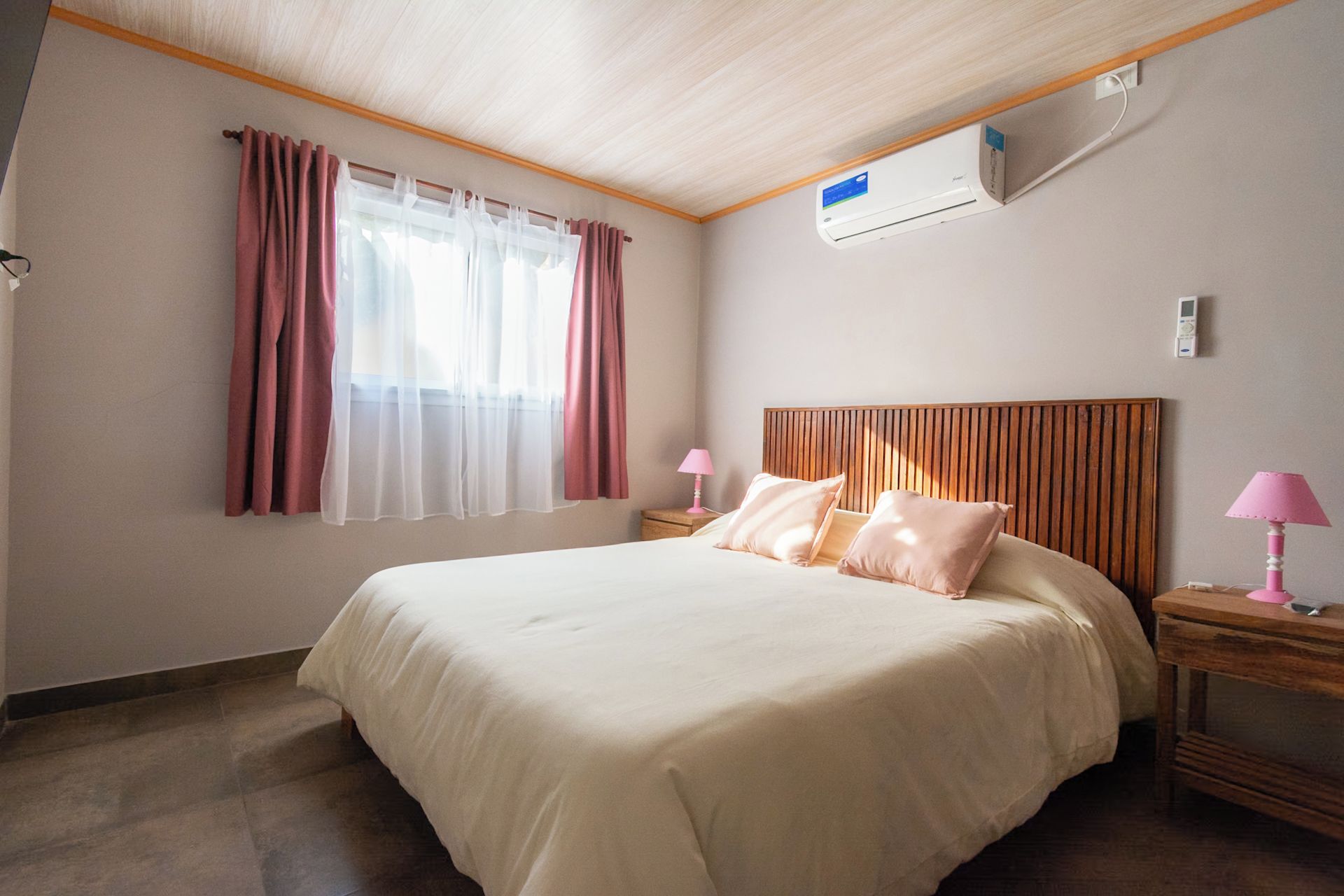 Habitación de hotel con cama, dos mesitas de noche, ventana con cortinas, unidad de aire acondicionado y techo de madera.