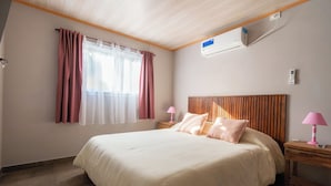 Quarto de hotel com cama, duas mesinhas de cabeceira, janela com cortinas, aparelho de ar condicionado e teto de madeira.