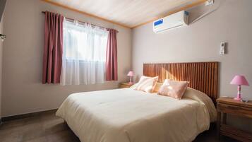 Habitación de hotel con cama, dos mesitas de noche, ventana con cortinas, unidad de aire acondicionado y techo de madera.