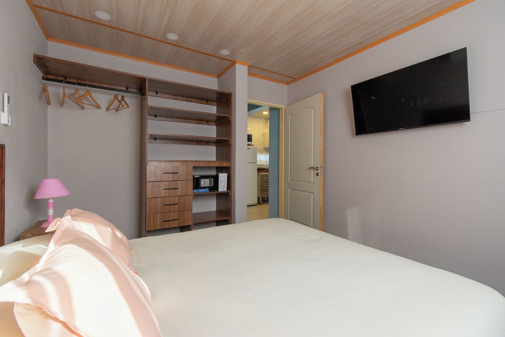 Un dormitorio con cama, televisor montado en la pared, armario con varios compartimentos y una puerta que da a otra habitación.