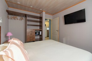 Un dormitorio con cama, televisor montado en la pared, armario con varios compartimentos y una puerta que da a otra habitación.