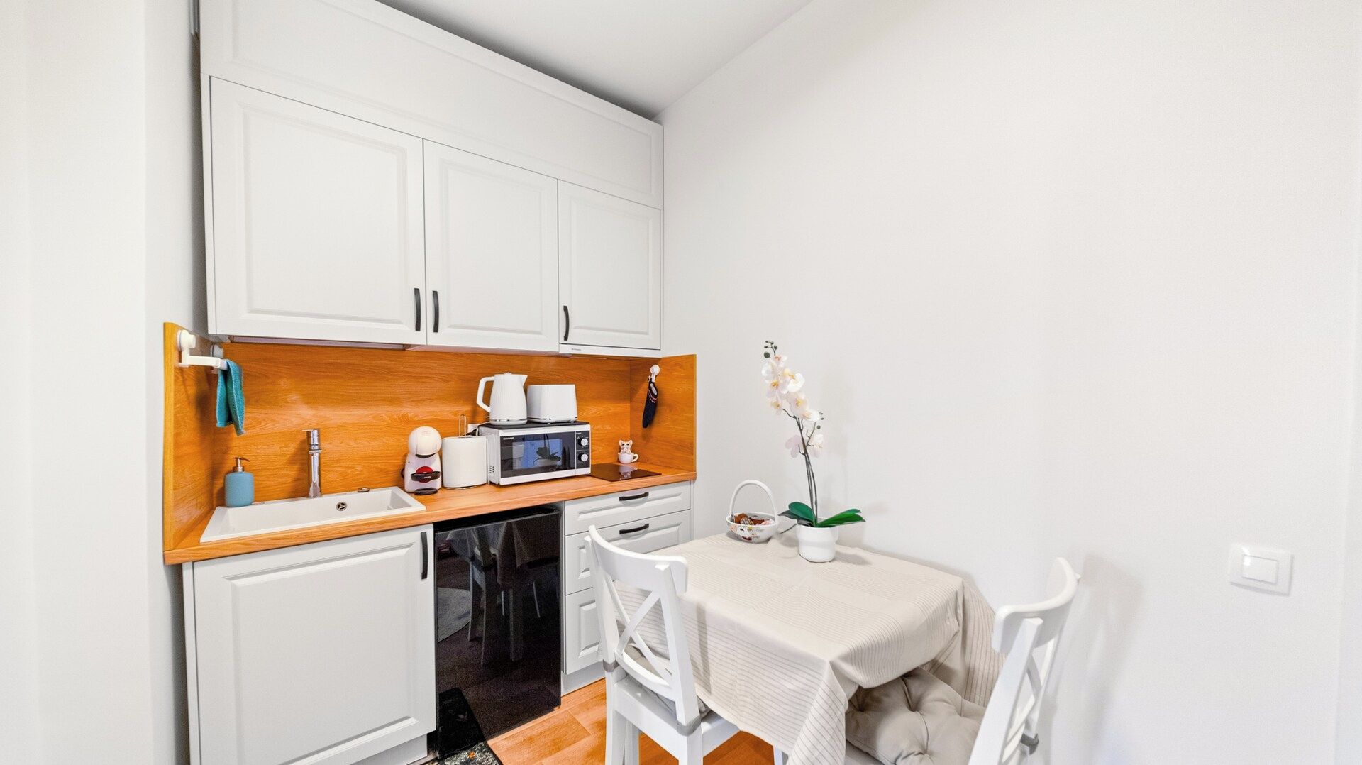 A compact kitchenette with white cabinets, a wooden countertop, a microwave, a kettle, and a small table with chairs.