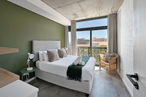 Ein Hotelzimmer mit Bett, Nachttisch, Lampe und Blick auf die Stadt durch große Fenster.