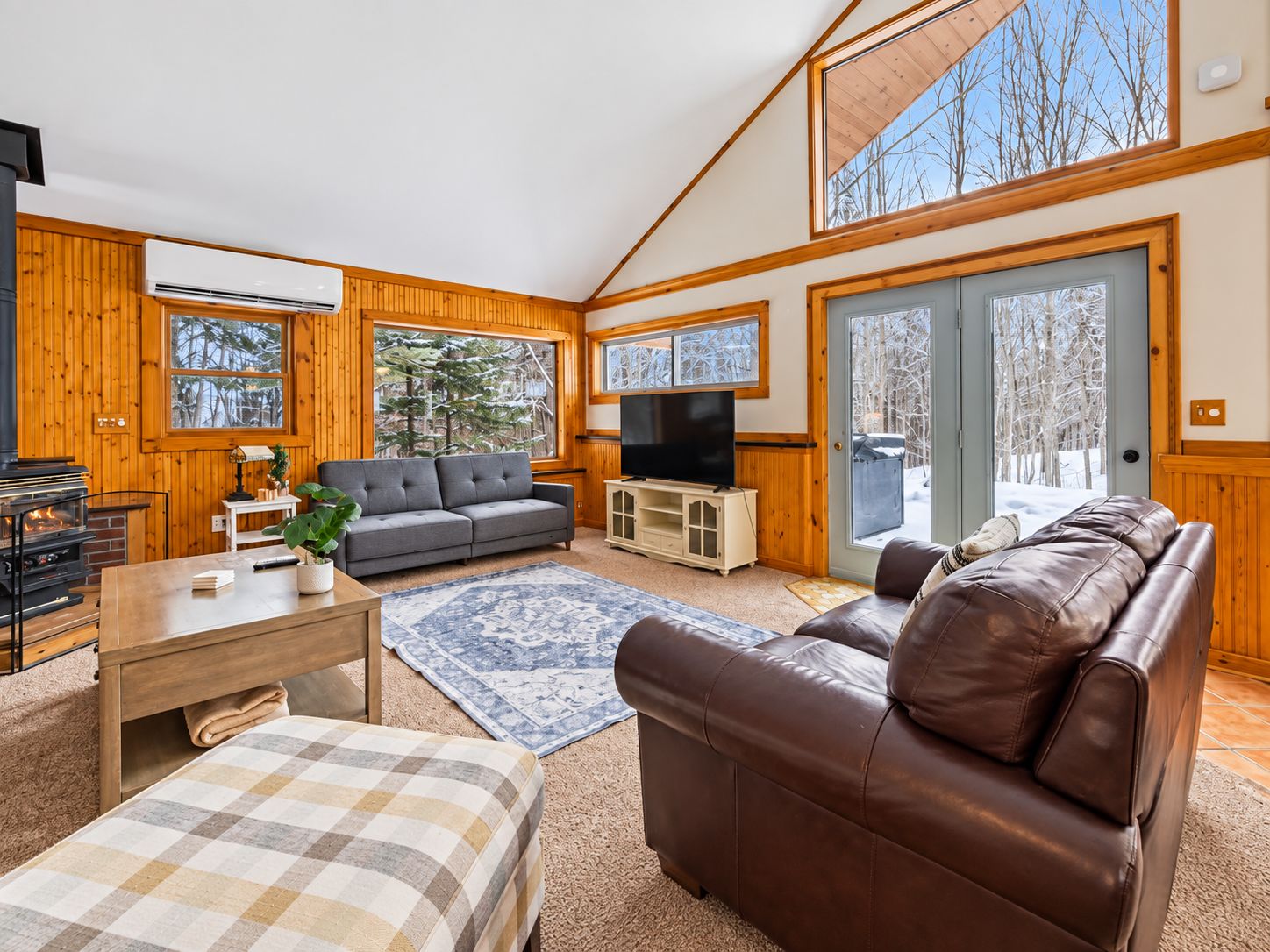 A cozy living room with a fireplace, a sofa, a television, and a large window offering a view of trees.