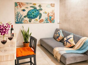 Quarto com sofá cinzento, cadeira de madeira com almofada laranja, mesa com copos de vinho e garrafa, planta em vaso e pintura de uma tartaruga e peixes na parede.