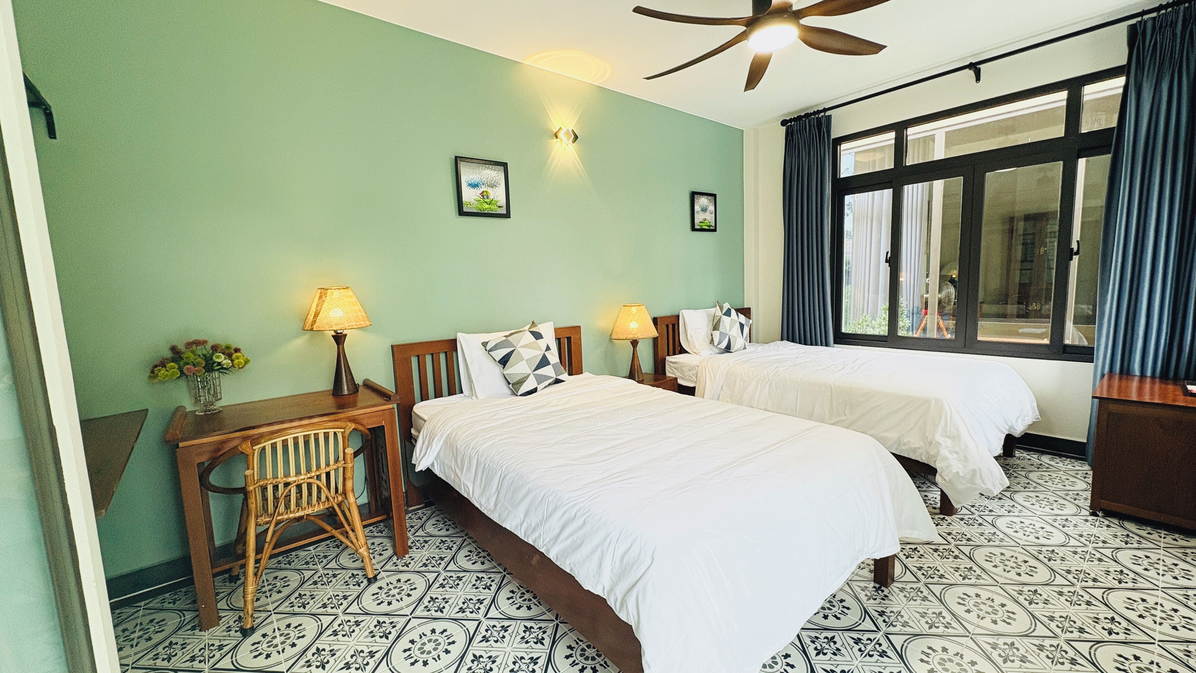 Une chambre d’hôtel avec deux lits, un bureau, une chaise, un ventilateur de plafond et une fenêtre avec des rideaux.