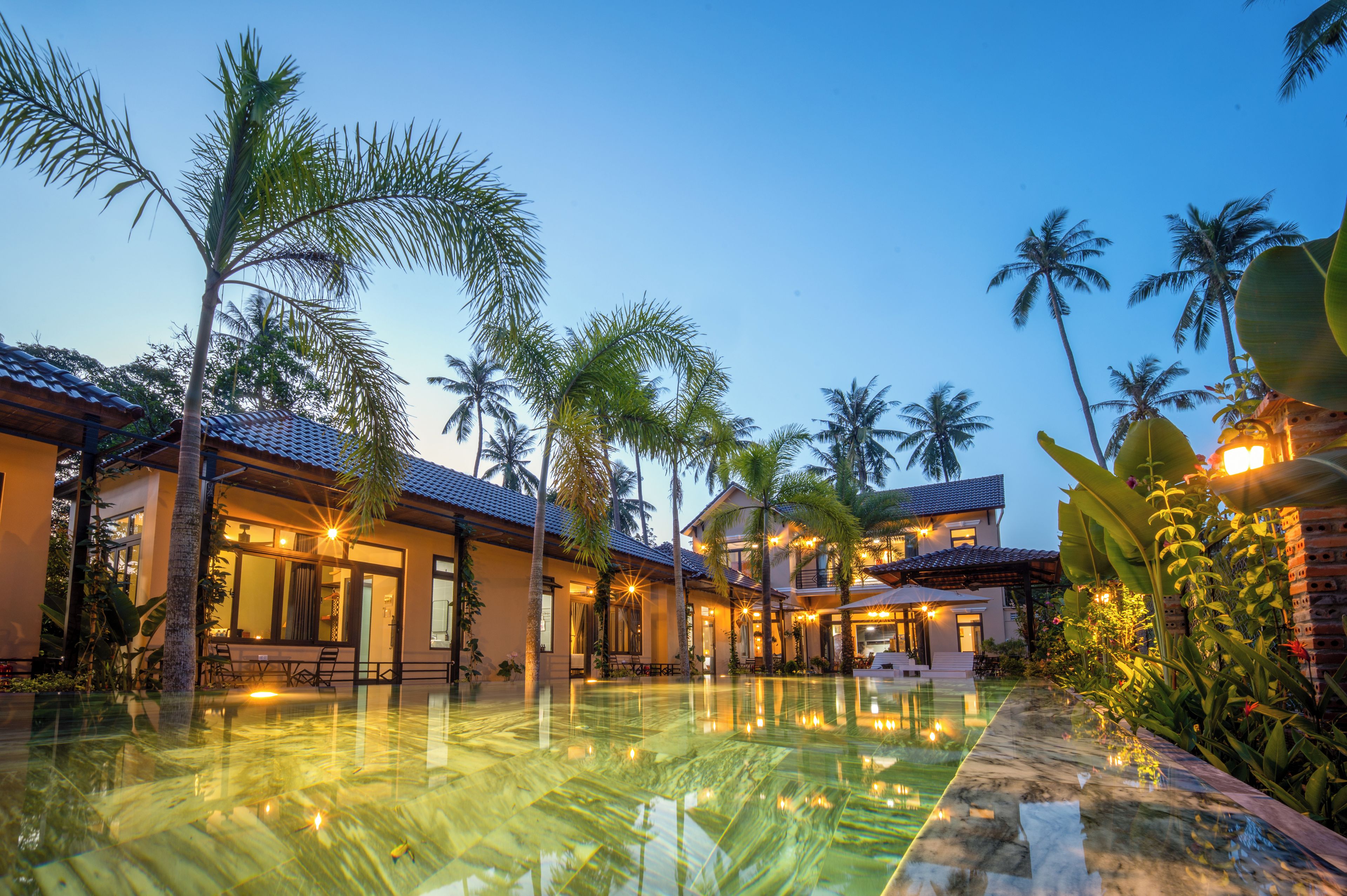 A resort with a swimming pool, palm trees, and multiple buildings with warm lighting.