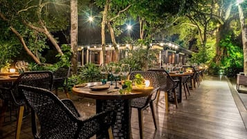 Un área de comedor al aire libre con sillas y mesas de mimbre, rodeada de árboles e iluminada por luces colgantes.